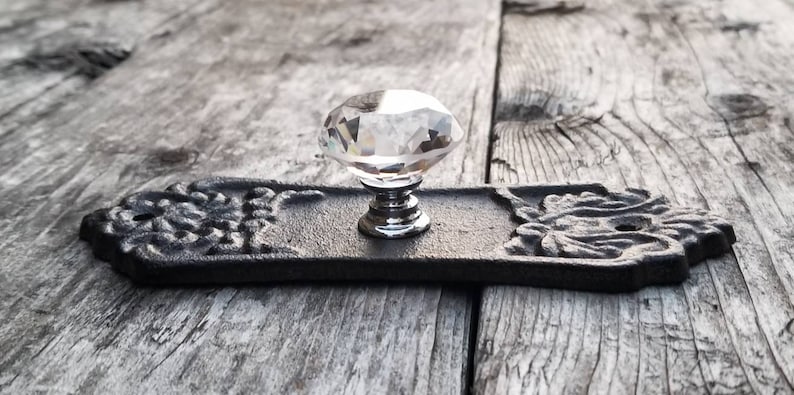 May include: A decorative cabinet pull with a clear crystal knob and a black, ornate backplate. The backplate features floral designs on each end. The hardware is designed for furniture and home decor.