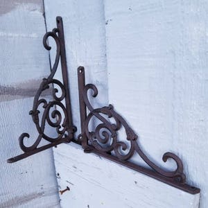 Puede incluir: Dos soportes de estante de metal adornados con un dise&ntilde;o de desplazamiento. Los soportes est&aacute;n hechos de metal marr&oacute;n oscuro y tienen un acabado r&uacute;stico.