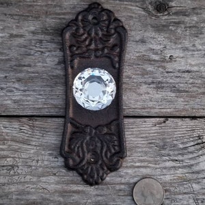 May include: Ornate, dark brown cast iron door pull with a large, clear crystal knob. The pull has decorative floral and scrollwork details. A quarter is next to the pull for size comparison. The door pull is on a weathered wooden surface.