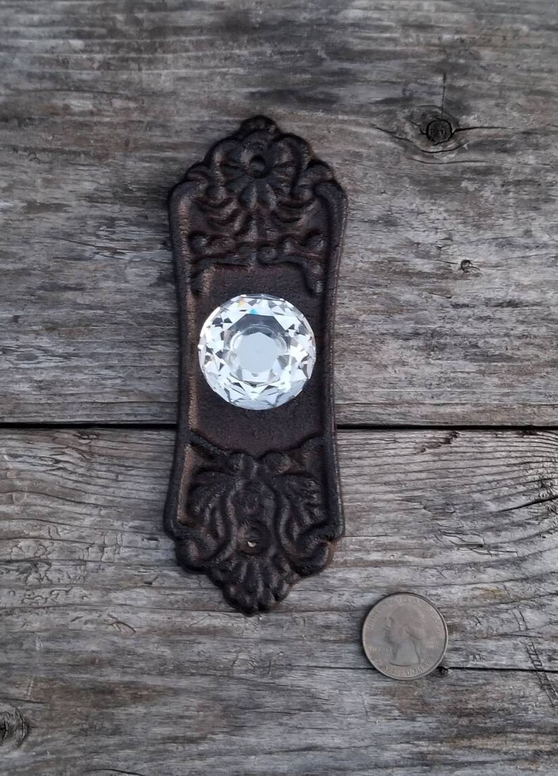 May include: Ornate, dark brown cast iron door pull with a large, clear crystal knob. The pull has decorative floral and scrollwork details. A quarter is next to the pull for size comparison. The door pull is on a weathered wooden surface.