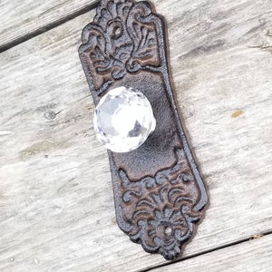 May include: Ornate, dark brown cast iron door pull with a clear crystal knob. The rectangular backplate features a detailed floral and scroll design. A vintage-style hardware piece.