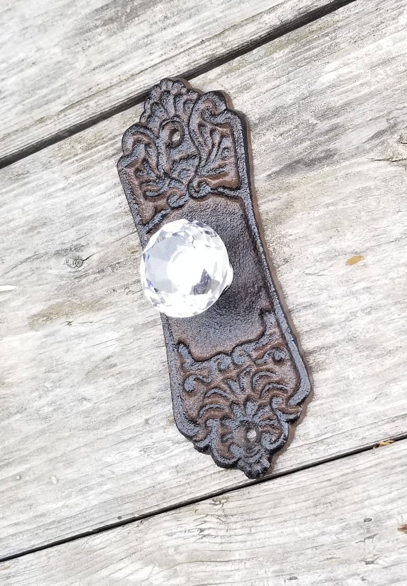 May include: Ornate, dark brown cast iron door pull with a clear crystal knob. The rectangular backplate features a detailed floral and scroll design. A vintage-style hardware piece.