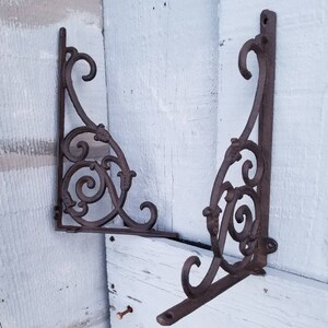 Puede incluir: Dos soportes de estante de hierro fundido adornados con un dise&ntilde;o de desplazamiento. Los soportes son de color marr&oacute;n oscuro y tienen un acabado r&uacute;stico.
