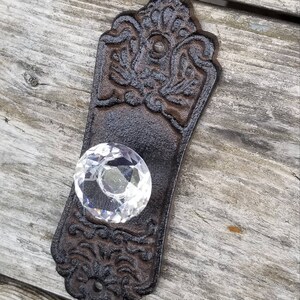 May include: Ornate, dark brown cast iron door pull with a clear, faceted crystal knob. The pull has a decorative floral and scroll design. A vintage-style hardware piece.