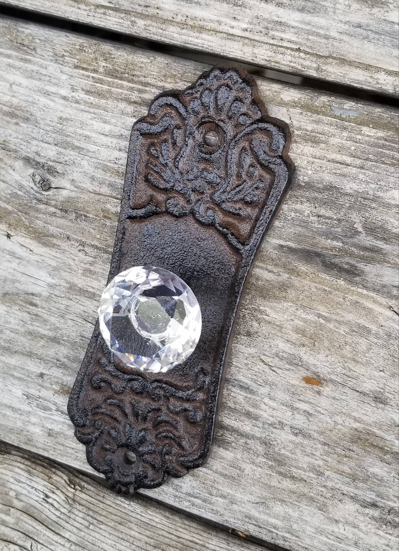 May include: Ornate, dark brown cast iron door pull with a clear, faceted crystal knob. The pull has a decorative floral and scroll design. A vintage-style hardware piece.