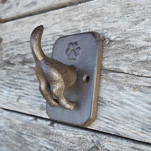May include: A dark brown cast iron wall hook featuring the rear end of a dog with an upright tail. The rectangular mounting plate includes a single screw hole and an embossed paw print design above the dog figure, providing a functional hook.