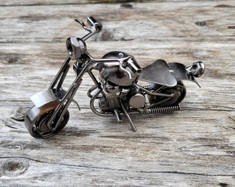 May include: A handcrafted metal motorcycle sculpture. The miniature motorcycle is silver-colored and features detailed construction, including a seat, handlebars, and wheels. The sculpture is displayed on a weathered wooden surface.