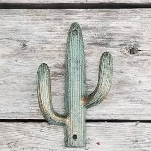 May include: A cast metal cactus-shaped wall hook with a distressed turquoise and gold finish. The hook has a vertical main stem with two curved arms extending outwards. The hook is mounted on a weathered wooden surface.