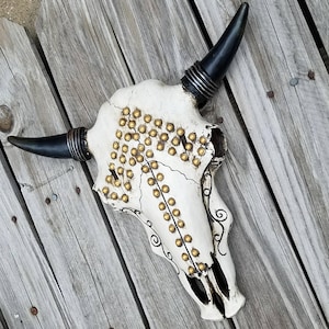 Peut inclure: Cr&acirc;ne de taureau blanc avec des cornes noires et des clous dor&eacute;s. Le cr&acirc;ne est d&eacute;cor&eacute; d'un motif stylis&eacute; et a un aspect vieilli.