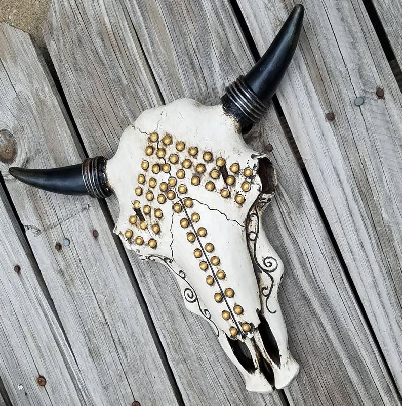 Peut inclure: Cr&acirc;ne de taureau blanc avec des cornes noires et des clous dor&eacute;s. Le cr&acirc;ne est d&eacute;cor&eacute; d'un motif stylis&eacute; et a un aspect vieilli.