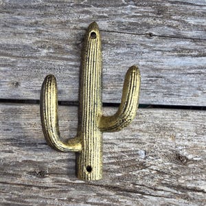 May include: A gold-colored metal cactus-shaped wall hook. The hook has a textured surface with vertical lines and two curved arms extending from the main body. The hook is designed to be mounted on a wall and has two holes for screws.