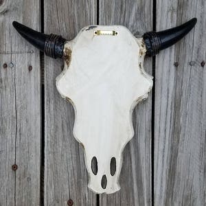 Peut inclure: Cr&acirc;ne de taureau en fausse taxidermie blanc avec des cornes noires et un support en bois. Le cr&acirc;ne a une finition blanche vieillie et des accents noirs autour des cornes.