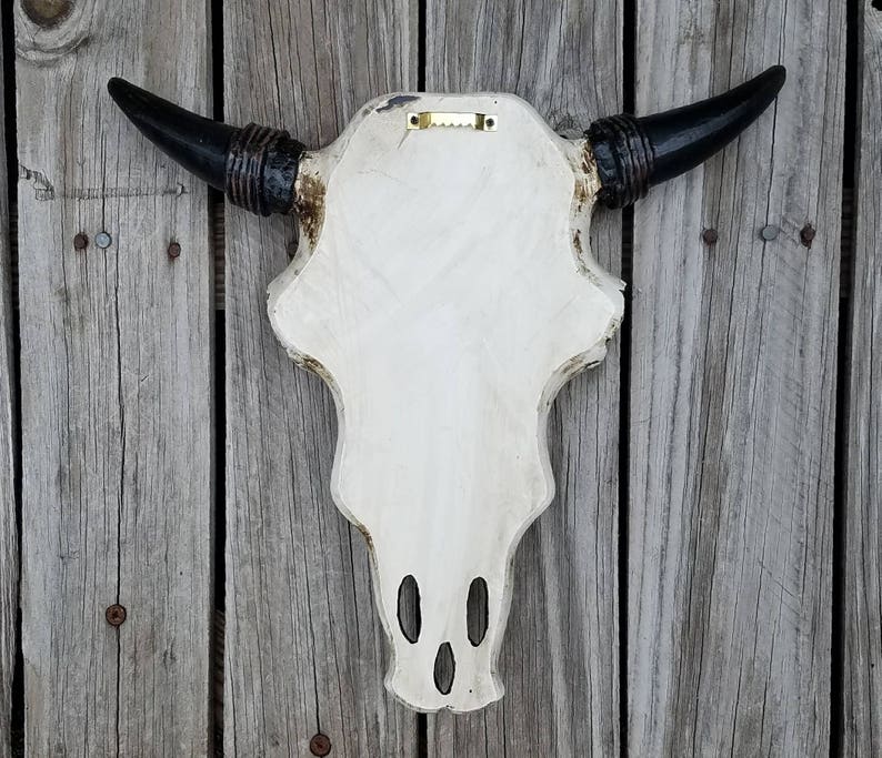 Peut inclure: Cr&acirc;ne de taureau en fausse taxidermie blanc avec des cornes noires et un support en bois. Le cr&acirc;ne a une finition blanche vieillie et des accents noirs autour des cornes.