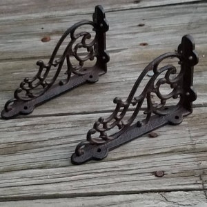 May include: Two ornate, dark brown cast iron shelf brackets. Each bracket features a decorative scroll design and mounting holes. The brackets are displayed on a weathered wooden surface, showcasing their rustic charm.