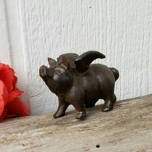K&ouml;nnte beinhalten: Eine Schweinefigur aus Gusseisen mit Fl&uuml;geln. Das Schwein ist braun und hat ein detailliertes Gesicht. Es steht auf einer Holzoberfl&auml;che.
