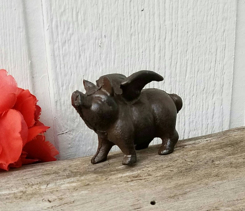 K&ouml;nnte beinhalten: Eine Schweinefigur aus Gusseisen mit Fl&uuml;geln. Das Schwein ist braun und hat ein detailliertes Gesicht. Es steht auf einer Holzoberfl&auml;che.
