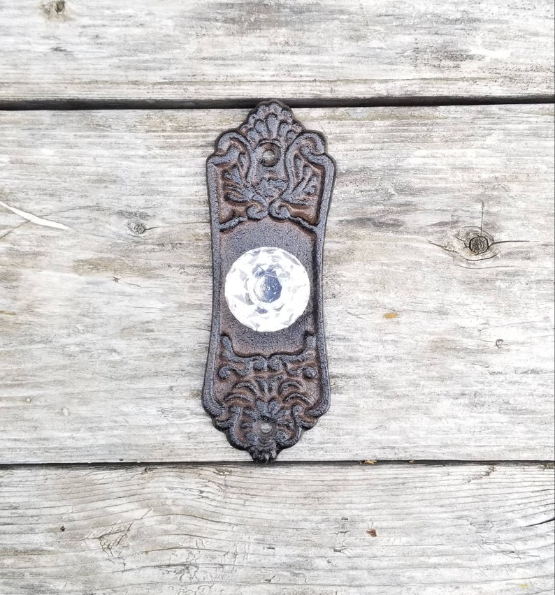 May include: Ornate, antique-style door knob with a clear, faceted glass knob. The dark brown metal backplate features detailed floral and scrollwork designs. The knob is centered on a weathered wooden surface.