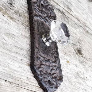May include: A decorative door handle with a clear crystal knob and a dark brown, ornate backplate. The backplate has a detailed, textured design. The handle is mounted on a light-colored wooden surface.