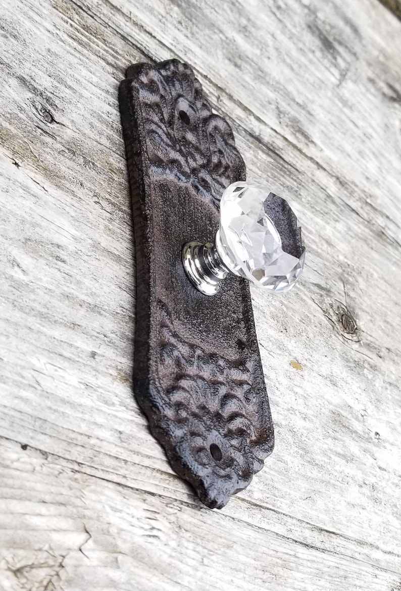 May include: A decorative door handle with a clear crystal knob and a dark brown, ornate backplate. The backplate has a detailed, textured design. The handle is mounted on a light-colored wooden surface.