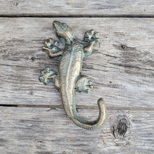 May include: A cast iron gecko hook with a green and gold finish. The hook is shaped like a gecko with its tail curled up. It is a decorative and functional piece that can be used to hang keys, towels, or other items.