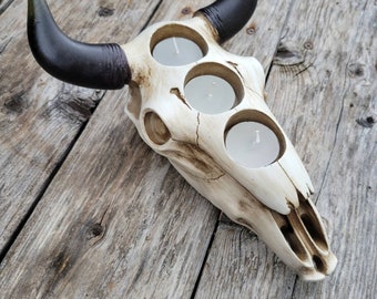 May include: A white ceramic bull skull candle holder with black horns. The skull has three circular cutouts for tealight candles.