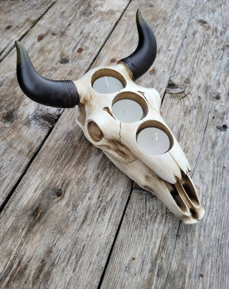 May include: A white ceramic bull skull candle holder with black horns. The skull has three circular cutouts for tealight candles.