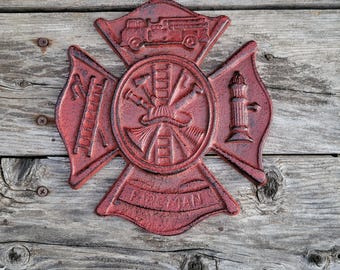 May include: A red cast iron firefighter emblem with a cross design. The emblem features a fire truck, ladder, and lighthouse. The word "FIREMAN" is inscribed on the bottom. The emblem is mounted on a weathered wooden surface.
