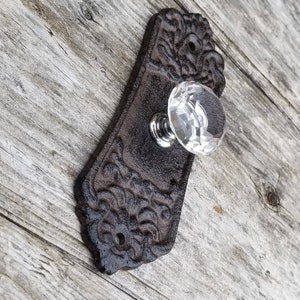 May include: Ornate, dark brown cast iron door knob with a clear, faceted crystal knob. The backplate has a decorative, swirling pattern. The knob is mounted on a weathered, gray wooden surface.
