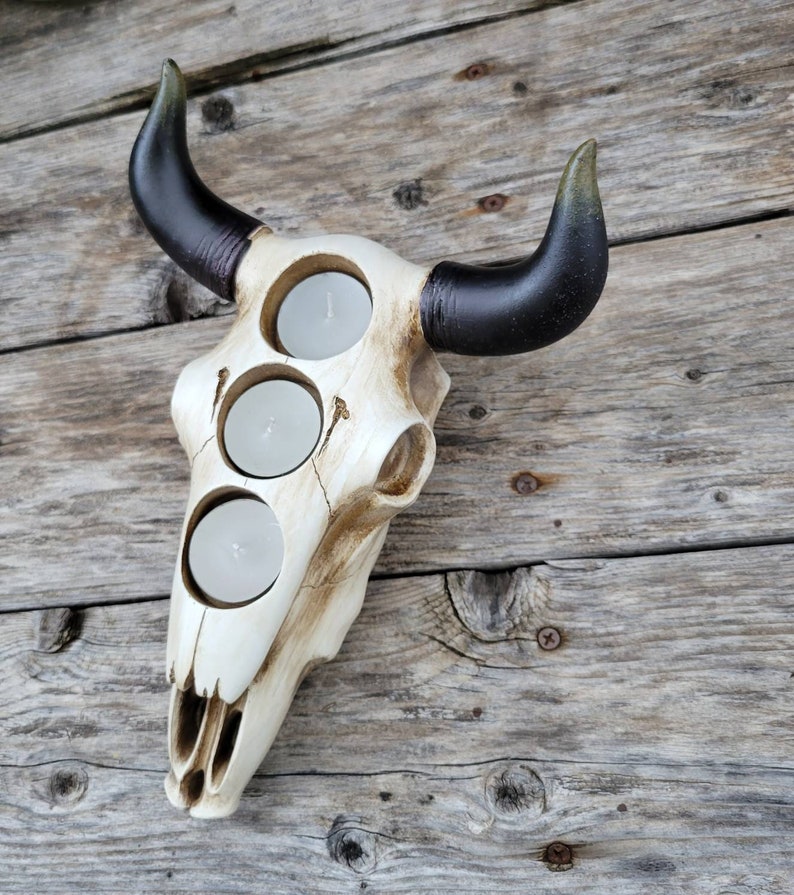 May include: White ceramic bull skull candle holder with three candle openings. The skull has a distressed, aged look and the horns are black with a green tip.