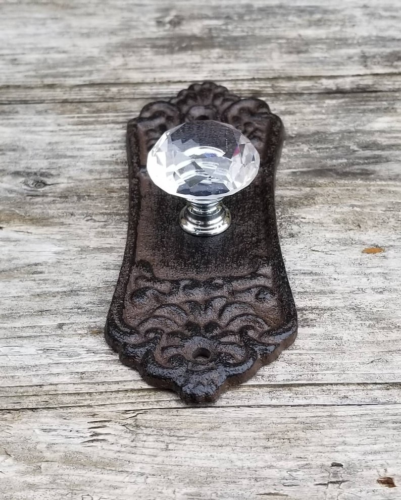 May include: A decorative, antique-style door knob with a clear, faceted crystal handle and a dark brown, ornate backplate. The backplate has a detailed, floral design. The knob is mounted on a weathered, wooden surface.