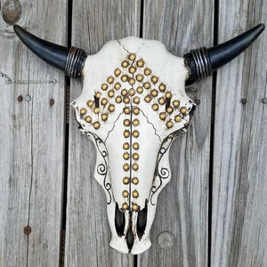 Peut inclure: Cr&acirc;ne de taureau blanc avec des clous dor&eacute;s et des cornes noires mont&eacute; sur un mur en bois.