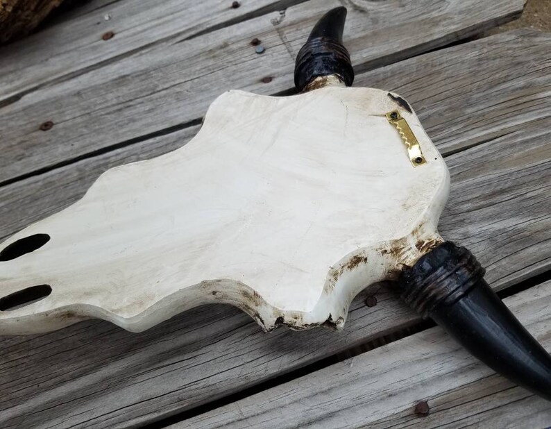 Peut inclure: D&eacute;coration murale en forme de cr&acirc;ne de taureau en bois blanc avec des cornes noires. Le cr&acirc;ne a une finition vieillie et un crochet de suspension en laiton.