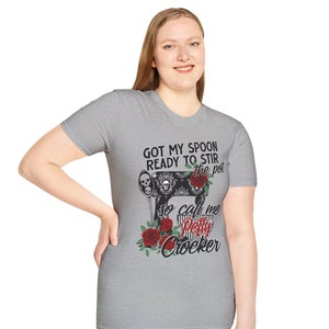 Peut inclure: Un t-shirt gris avec une illustration en noir et blanc d'une machine à coudre vintage avec des roses rouges et des crânes. Le texte sur le t-shirt est "Got my spoon ready to stir the pot so call me Petty Crocker".