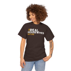 May include: A brown t-shirt with the text "THE REAL HOUSEWIVES OF UPS" in white and gold lettering. The shirt is being worn by a person with curly brown hair and blue jeans.