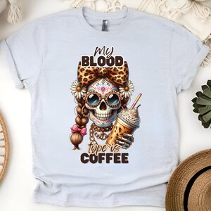 Puede incluir: Una camiseta blanca con un diseño de calavera de azúcar colorida sosteniendo una bebida de café. El texto en la camiseta dice "My Blood Type is Coffee".
