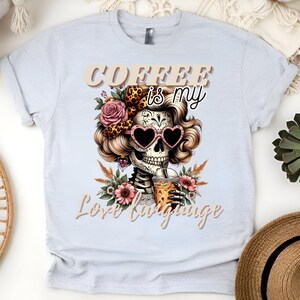 May include: A white t-shirt featuring a graphic of a sugar skull with flowers and a coffee cup. The text "Coffee is my love language" is printed on the shirt.