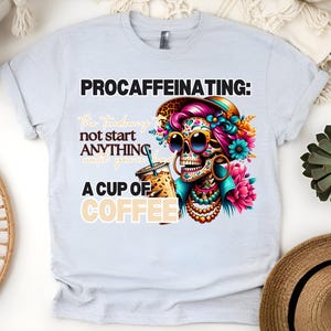May include: A light blue t-shirt featuring a colourful graphic of a sugar skull holding a cup of coffee. The text "Procaffeinating: The tendency to not start anything until you've had a cup of coffee" is printed on the shirt.