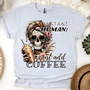 May include: White t-shirt featuring a graphic of a sugar skull with flowers and sunglasses holding a cup of coffee. The text "Instant Human: Just Add Coffee" is printed on the shirt.