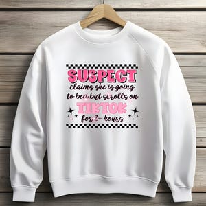 Puede incluir: Sudadera blanca con un borde de cuadros negros y blancos alrededor del texto "SUSPECT claims she is going to bed but scrolls on TIKTOK for 2+ hours". El texto está en rosa.