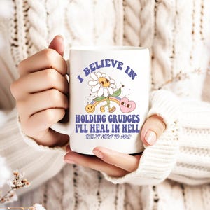 May include: White ceramic mug with a colorful cartoon daisy and rainbow design. The text reads "I believe in holding grudges I'll heal in hell right next to you!"