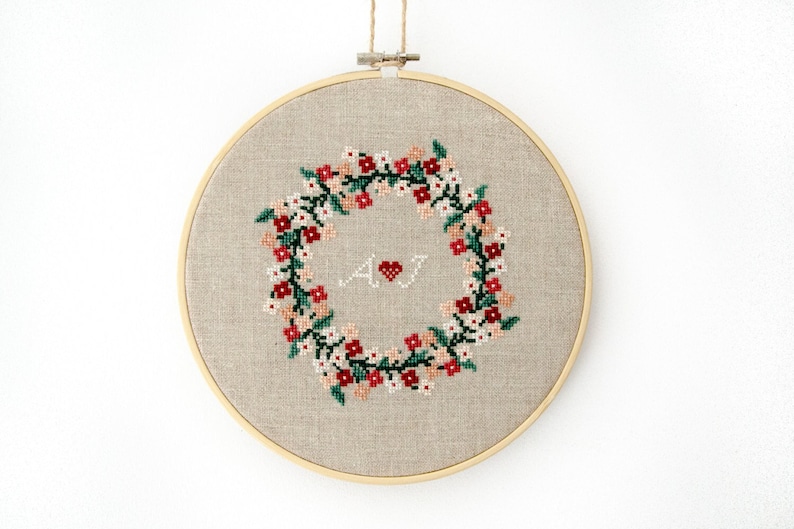 Puede incluir: Un aro de bordado beige con un dise&ntilde;o de punto de cruz de una corona de flores con flores rojas, rosas y blancas. Las iniciales "A" y "J" est&aacute;n en el centro de la corona con un peque&ntilde;o coraz&oacute;n rojo entre ellas.