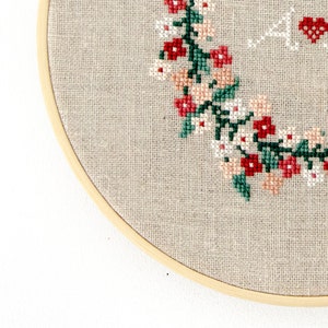 Puede incluir: Primer plano de un aro de bordado beige con un dise&ntilde;o floral cosido en hilo rojo, rosa y verde. El dise&ntilde;o presenta una corona de flores y hojas.