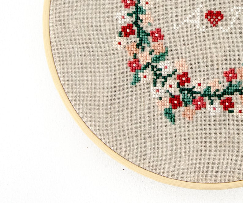 Puede incluir: Primer plano de un aro de bordado beige con un dise&ntilde;o floral cosido en hilo rojo, rosa y verde. El dise&ntilde;o presenta una corona de flores y hojas.