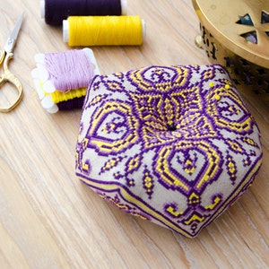 May include: A pentagon-shaped pincushion with a detailed purple and yellow embroidered design. Gold scissors, spools of purple and yellow thread, and a decorative brass container are arranged on a light wood surface, suggesting a sewing or craft theme.