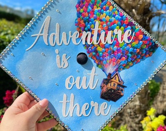 May include: A graduation cap decorated with a colorful hot air balloon design and the words "Adventure is out there." The cap has a blue background with rhinestone trim and the text "Fantasmil Art by Wensy".
