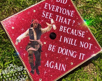 Puede incluir: Un gorro de graduación rojo con purpurina y ribete dorado. El gorro presenta una imagen del Capitán Jack Sparrow de las películas de Piratas del Caribe. El texto en el gorro dice "Did everyone see that? Because I will not be doing it again."
