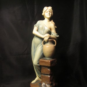 Könnte beinhalten: Eine Keramikstatue einer Frau in einem fließenden blauen Kleid, die einen braunen Tontopf hält. Die Statue steht auf einem braunen Steinsockel.