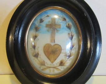 May include: Oval framed artwork featuring a gold cross above a gold heart, set against a blue and white background. The artwork is surrounded by a wreath of leaves and encased in a black oval frame.