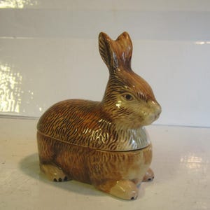 Może przedstawiać: Ceramiczna figurka królika z błyszczącym wykończeniem, przedstawiająca brązowo-beżowy wzór futra z teksturowanymi detalami. Królik jest przedstawiony w pozycji siedzącej z wyprostowanymi uszami, wyraźnym pyszczkiem i widocznymi łapkami.