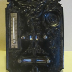 Puede incluir: Calendario de metal negro con detalles ornamentados, que incluye un termómetro con escalas Celsius y Fahrenheit, una pantalla de fecha y dos espacios en blanco para escribir. El termómetro marca 15 grados Celsius.